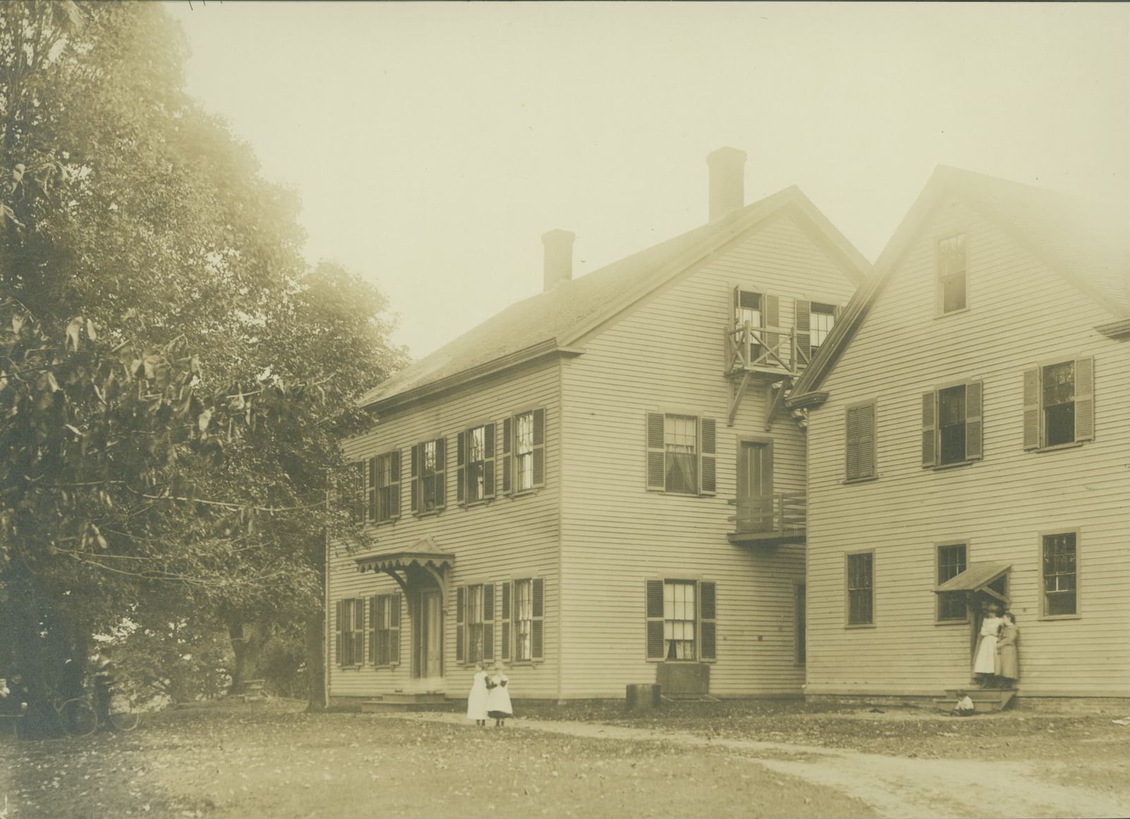 An archival photograph of a white clapboard building at Brook Farm, with children standing on the drive and trees around the house.