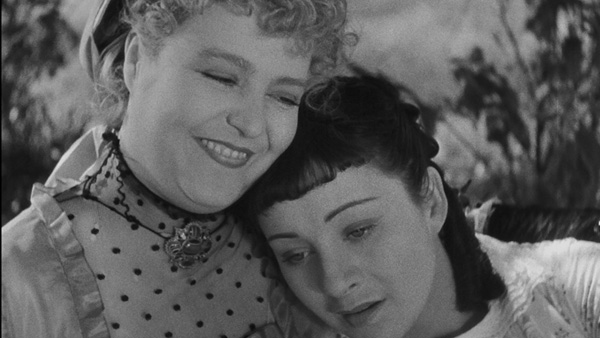 In a black-and-white film still, Henriette rests her head against her smiling mother's shoulder beneath leafy branches.