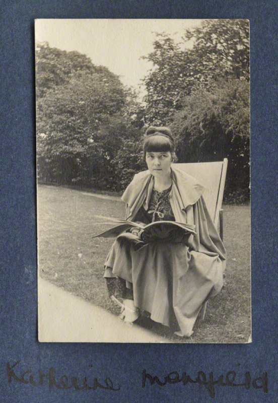 Katherine Mansfield seated in a garden chair with an open book across her lap, in a photograph mounted on a blue album page.