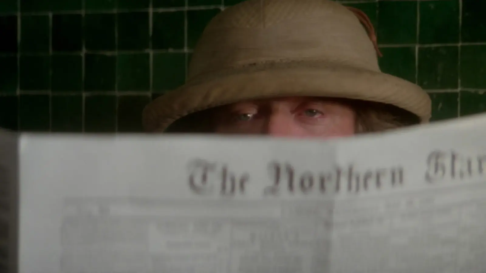 Michael Caine in a weathered pith helmet peers over the top of an open newspaper.
