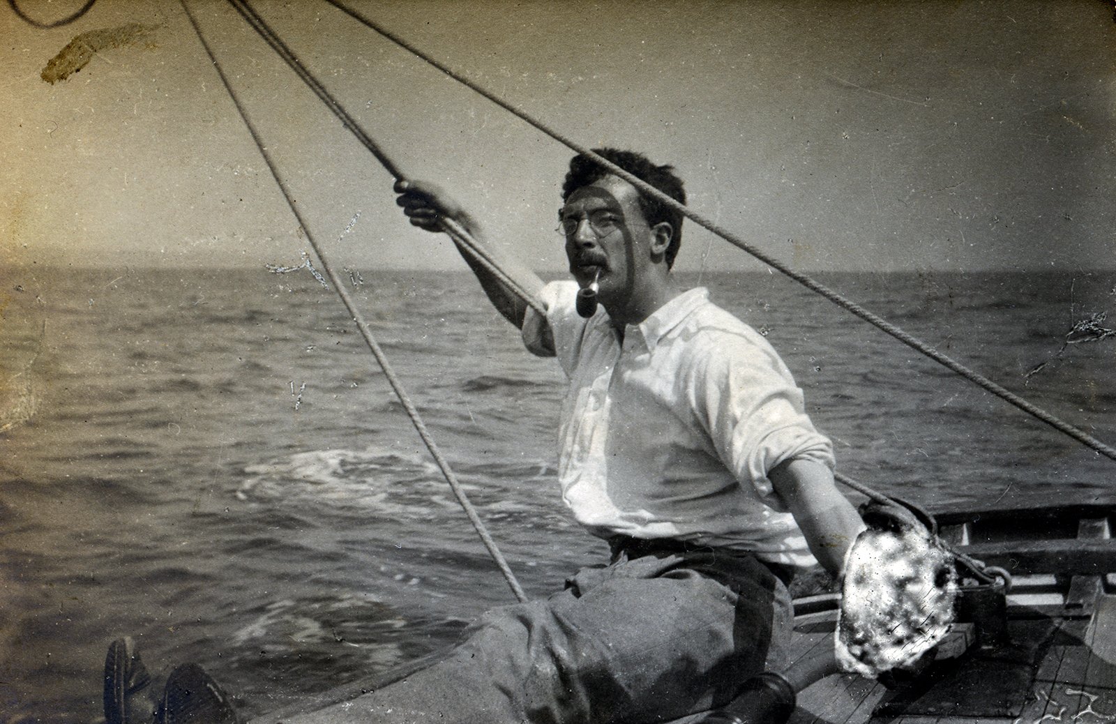Erskine Childers sits on Vixen's wooden deck, holding a rope and smoking a pipe, with open water behind him.