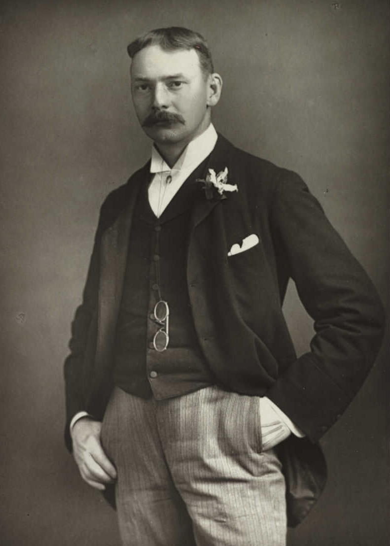 Jerome K. Jerome standing in a dark jacket and striped trousers, with one hand in his pocket, in an archival studio portrait.