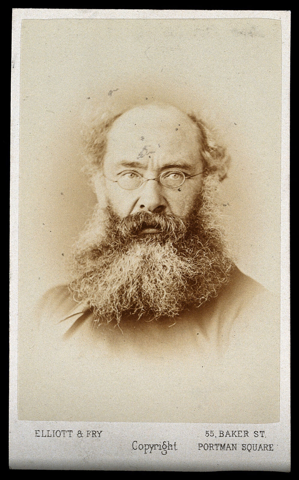 Archival studio photograph of Anthony Trollope, with a full beard and round spectacles, on an Elliott & Fry studio card.