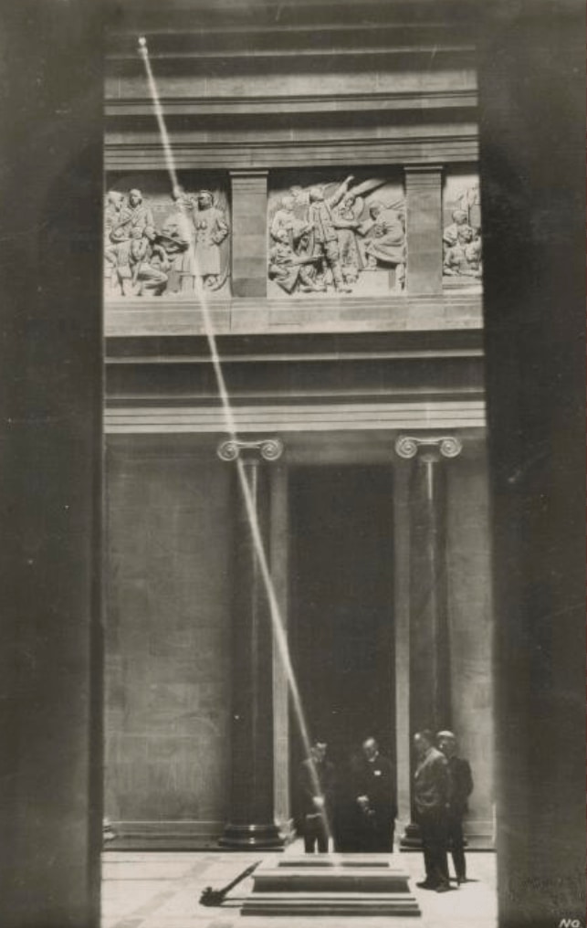 An archival black-and-white photograph of a beam of light crossing the Stone of Remembrance, with visitors and carved doorways behind it.