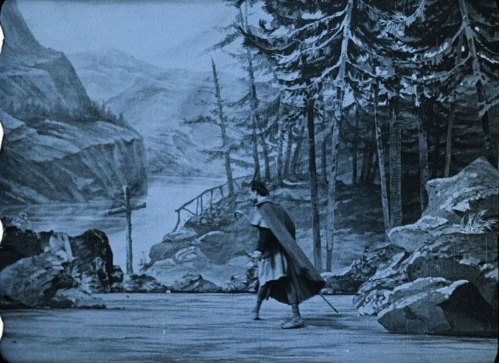 《威廉·退尔》（Guillaume Tell，1903）的一帧蓝色染色画面中，一个披着斗篷的人走过山间湖畔的岩石与树林布景。