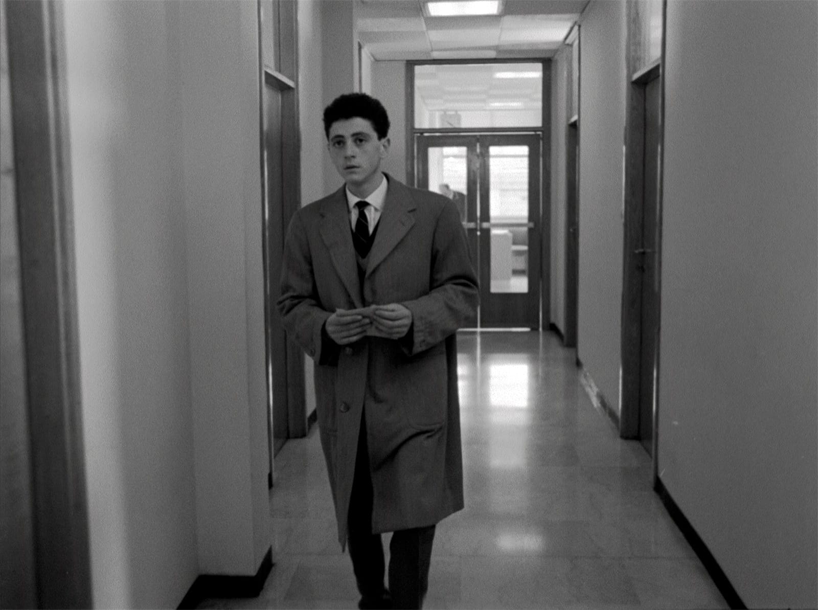 Sandro Panseri as Domenico stands in a long office corridor, wearing an overcoat and holding a small paper in both hands.