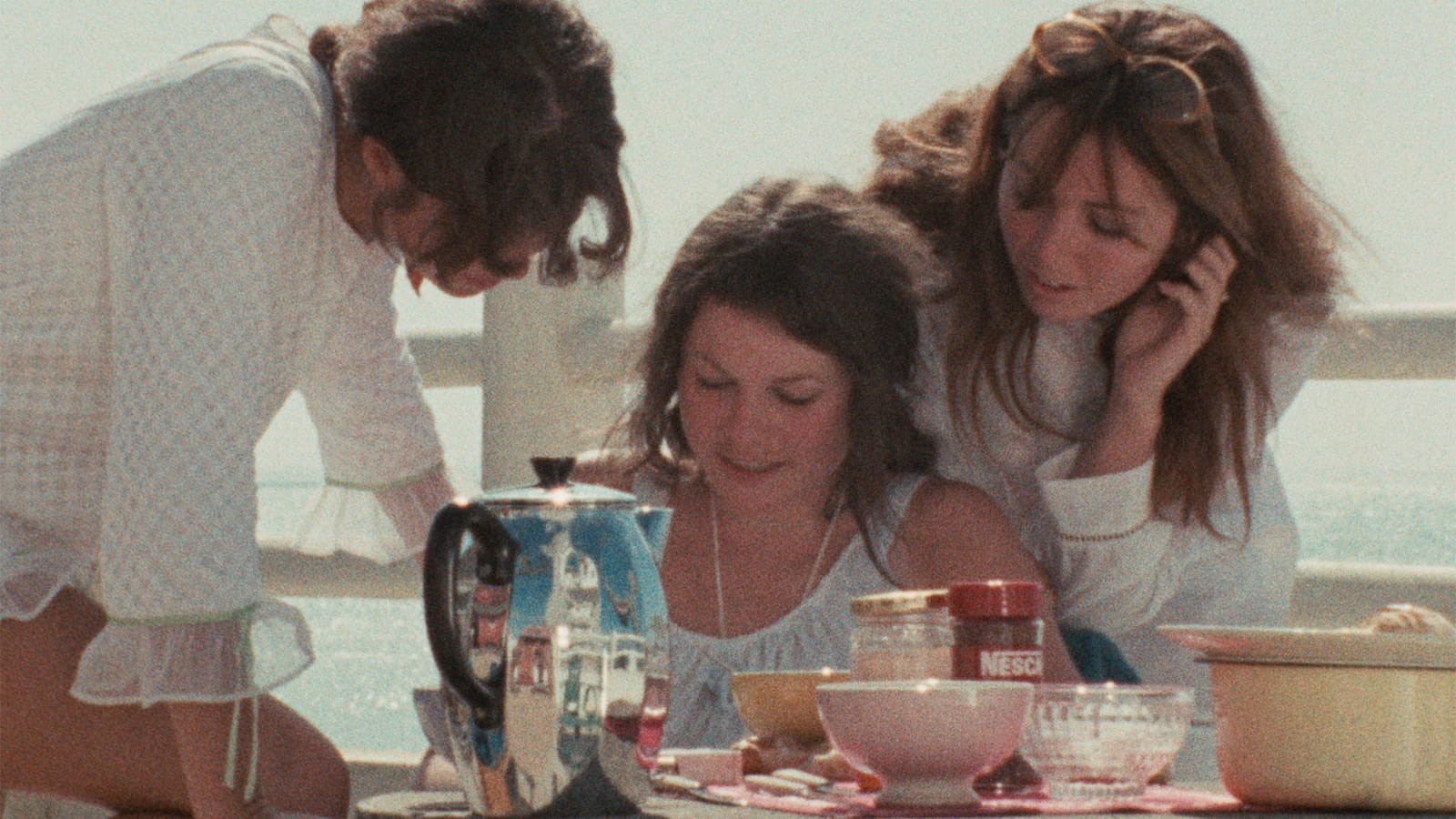 Three young women lean together over a breakfast table with bowls and a blue coffee pot in Near Orouët.
