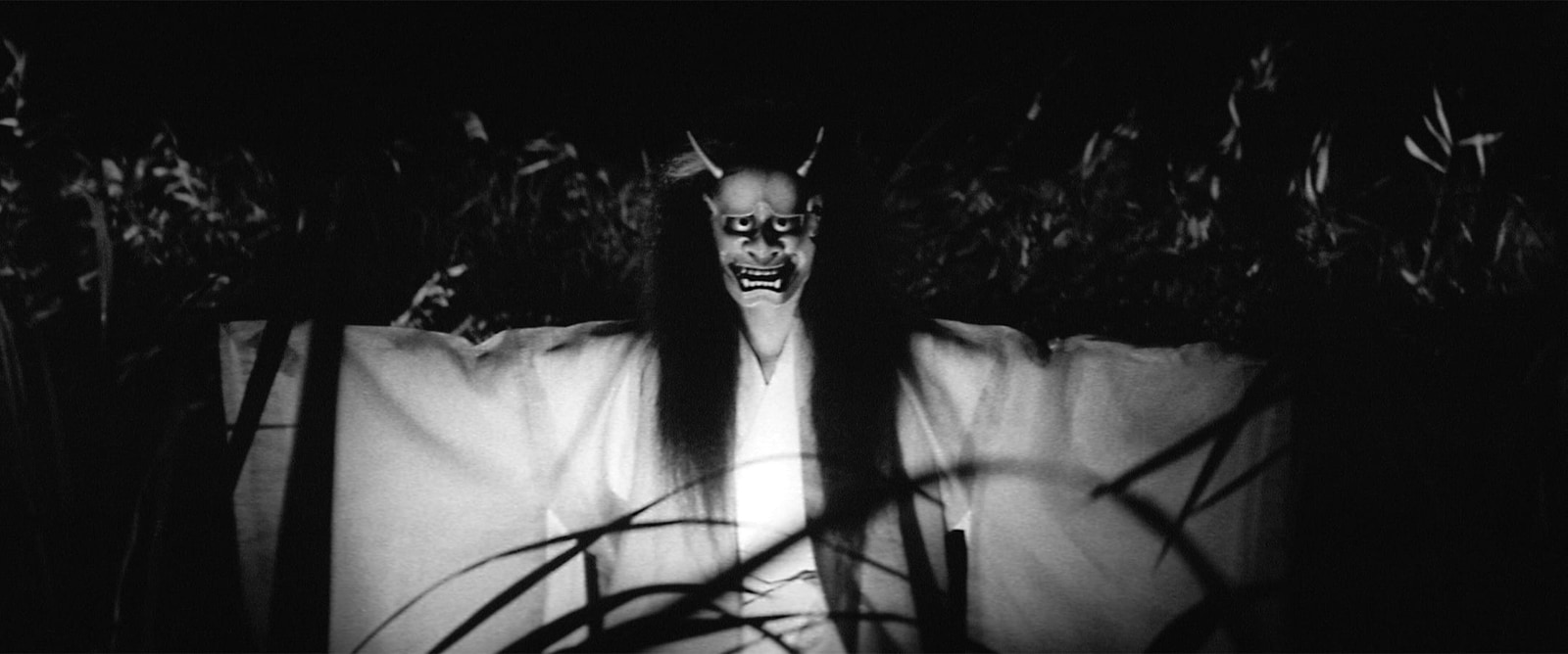 A masked figure spreads the sleeves of a pale robe among dark reeds, with stalks crossing the foreground.