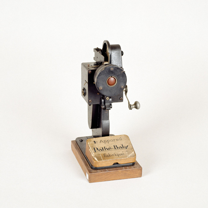 A surviving Pathé Baby prototype projector, with a hand crank and round front plate, mounted on a wooden base.