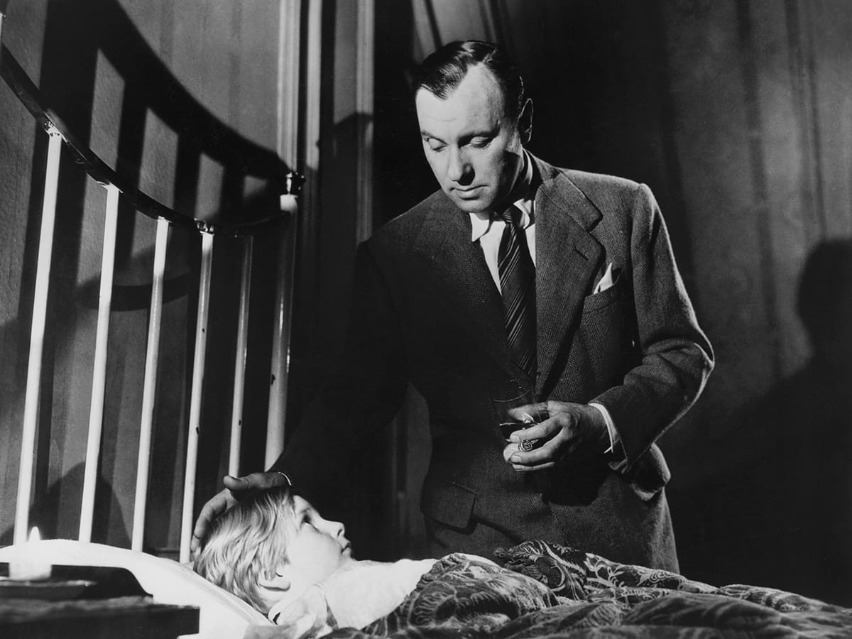 Ralph Richardson as Baines rests a hand on the head of Bobby Henrey's Philippe, who looks up from bed beside a metal headboard.