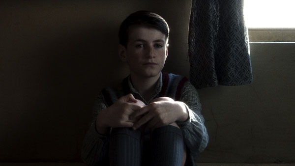 Leigh McCormack as Bud sits with his knees drawn up beside a bright window, his hands lit against the shadowed room.