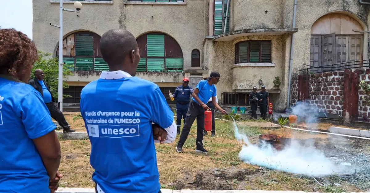 A workshop participant directs a fire extinguisher toward a smoking practice area while colleagues watch outside the Marien Ngouabi Museum.