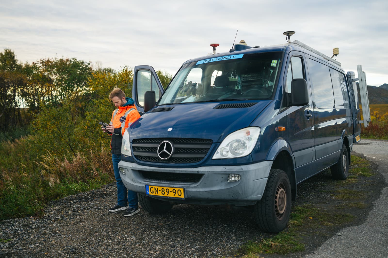 An ESA team member in an orange jacket operates a handheld device beside a blue navigation laboratory van fitted with roof antennas in Norway.