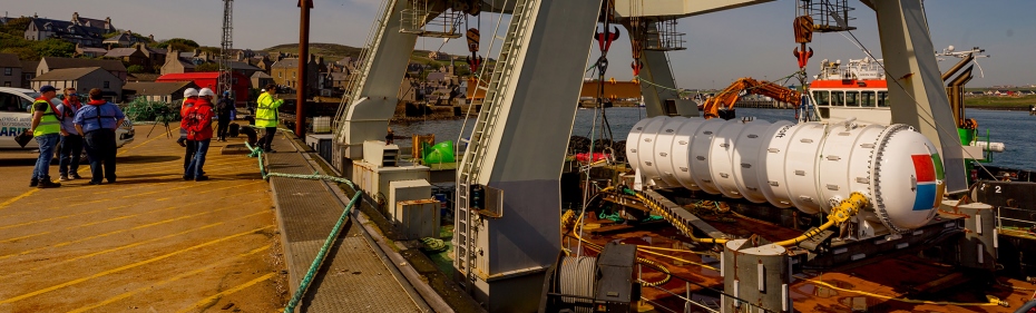 Crew members stand beside a gantry barge carrying the white cylindrical Northern Isles data centre in an Orkney harbour.