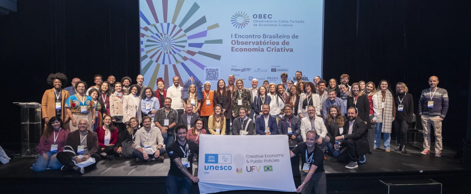 Participants pose together on stage beneath the banner of the Brazilian Meeting of Creative Economy Observatories in São Paulo.