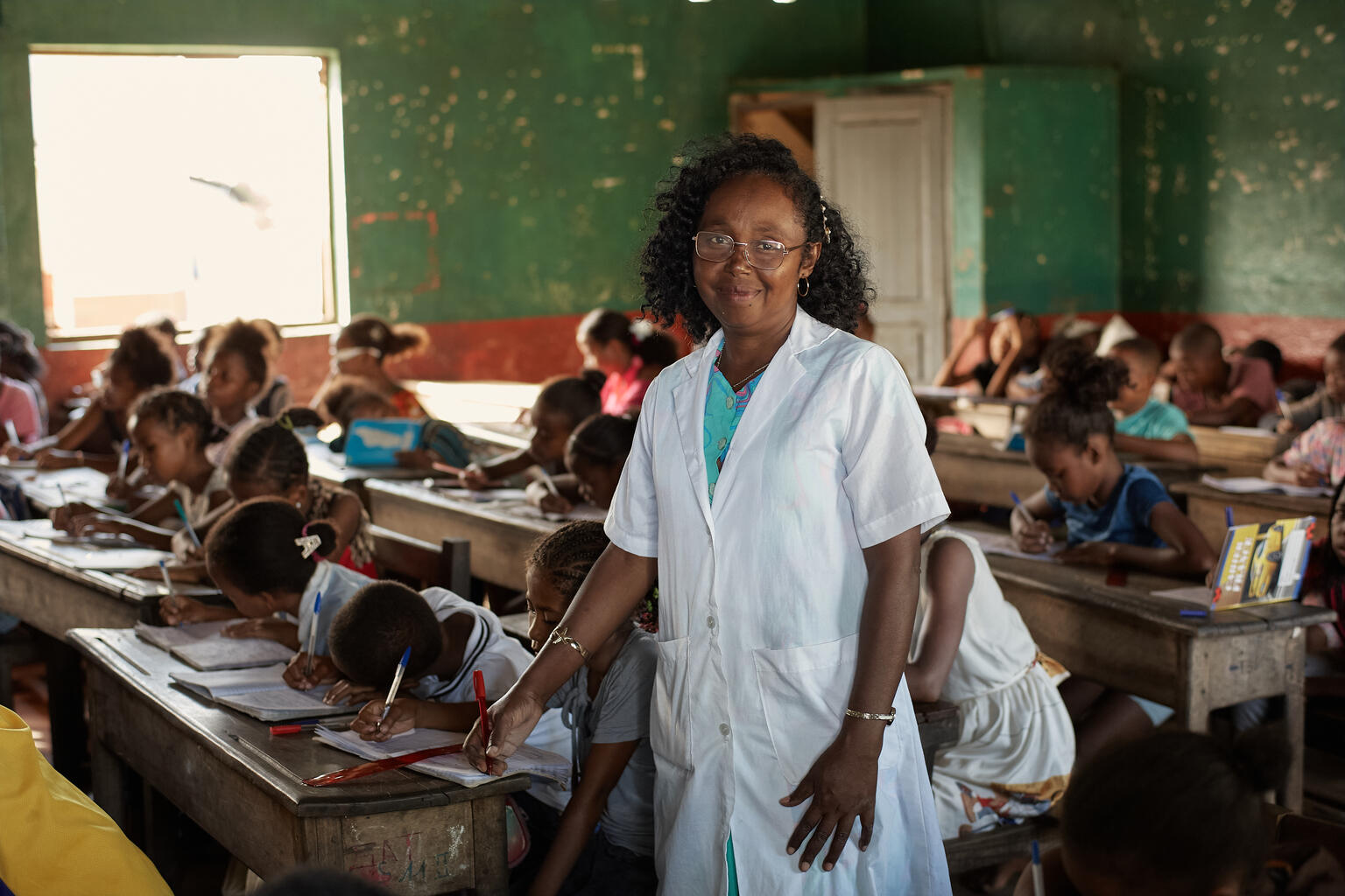 A teacher stands beside children writing at wooden desks in a classroom in Madagascar.