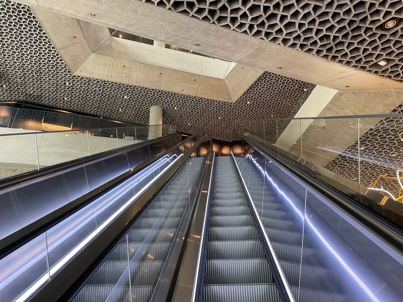 An escalator rises into Deichman Bjørvika beneath a honeycomb-patterned concrete ceiling and angular openings between floors.