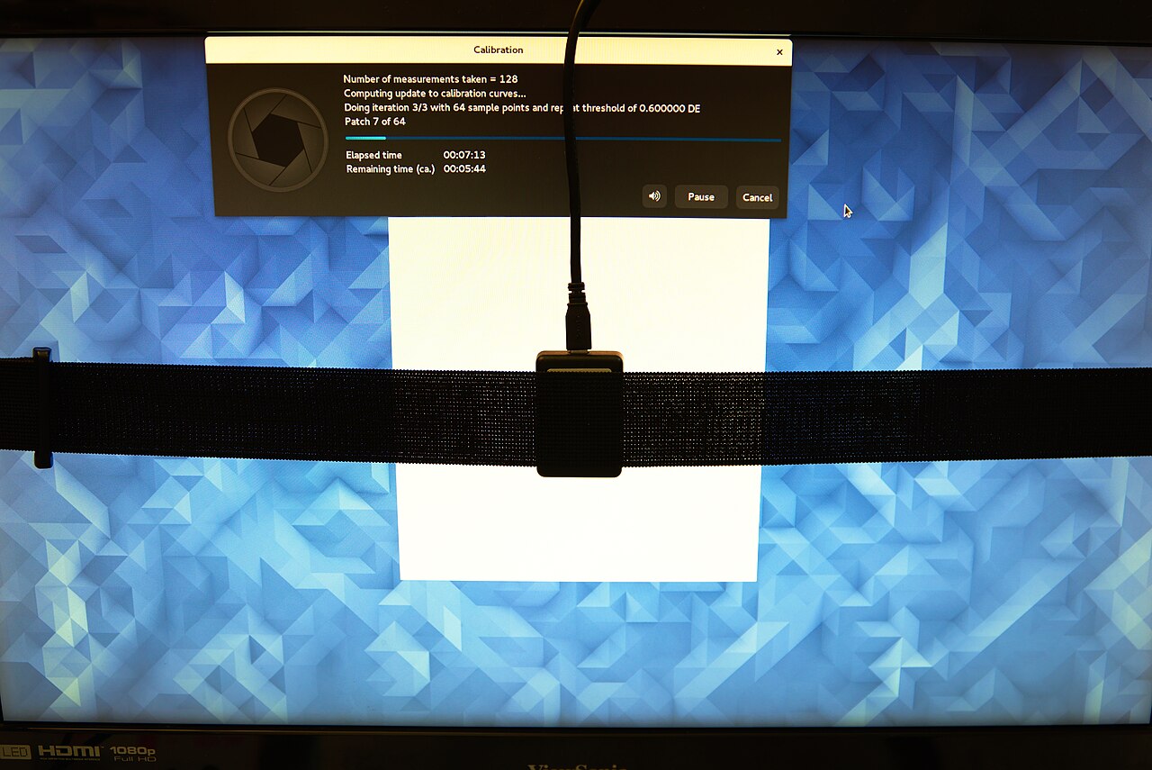 A black strap holds a ColorHug2 colorimeter against a white test patch on a monitor, with its cable hanging down from above.