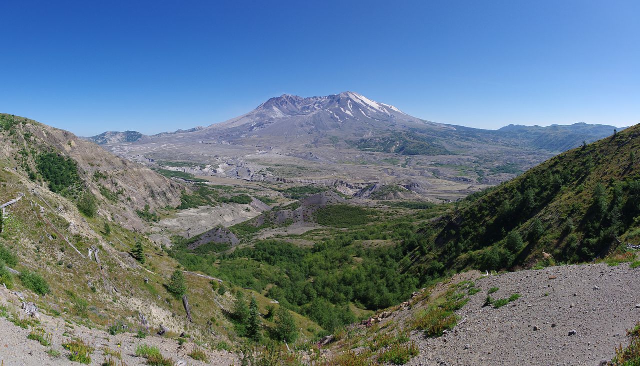 一幅拼接而成的风景照片：蓝天下，圣海伦斯火山耸立在绿色山脊与宽阔的浅色谷地之上。