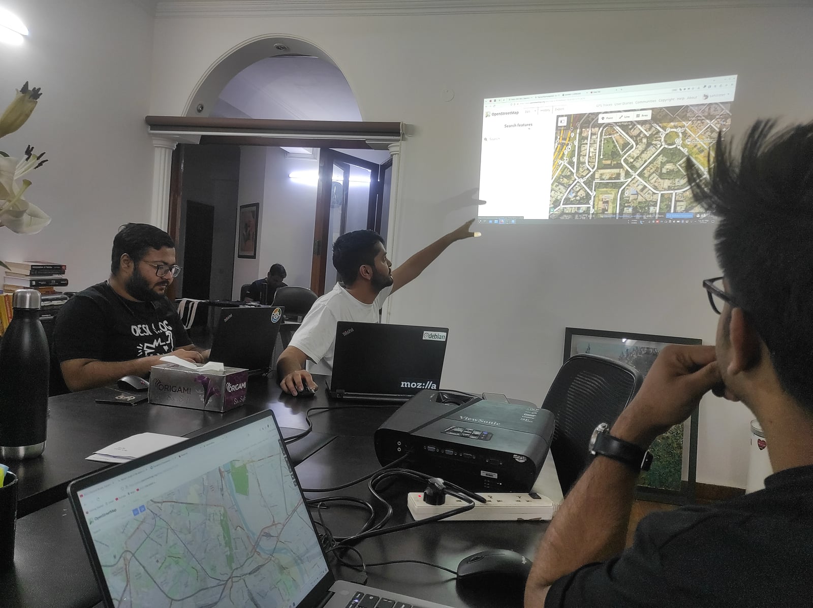 OpenStreetMap 工作坊的参与者围坐在笔记本电脑旁，一位讲师指向投影在墙上的航拍图像。