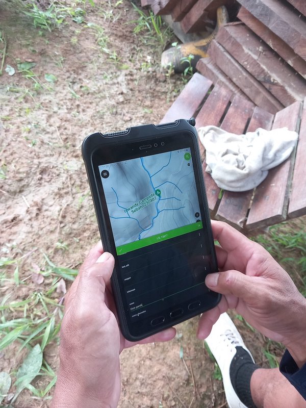 Hands hold a rugged mobile device displaying a field map beside a wooden bench and muddy ground.
