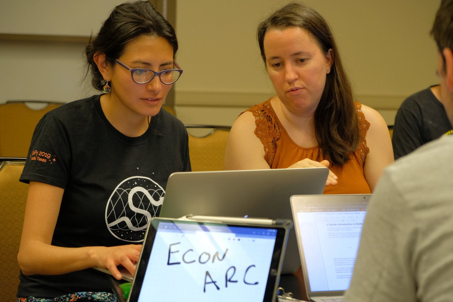 Two conference participants concentrate on a shared laptop at a table surrounded by other open computers.