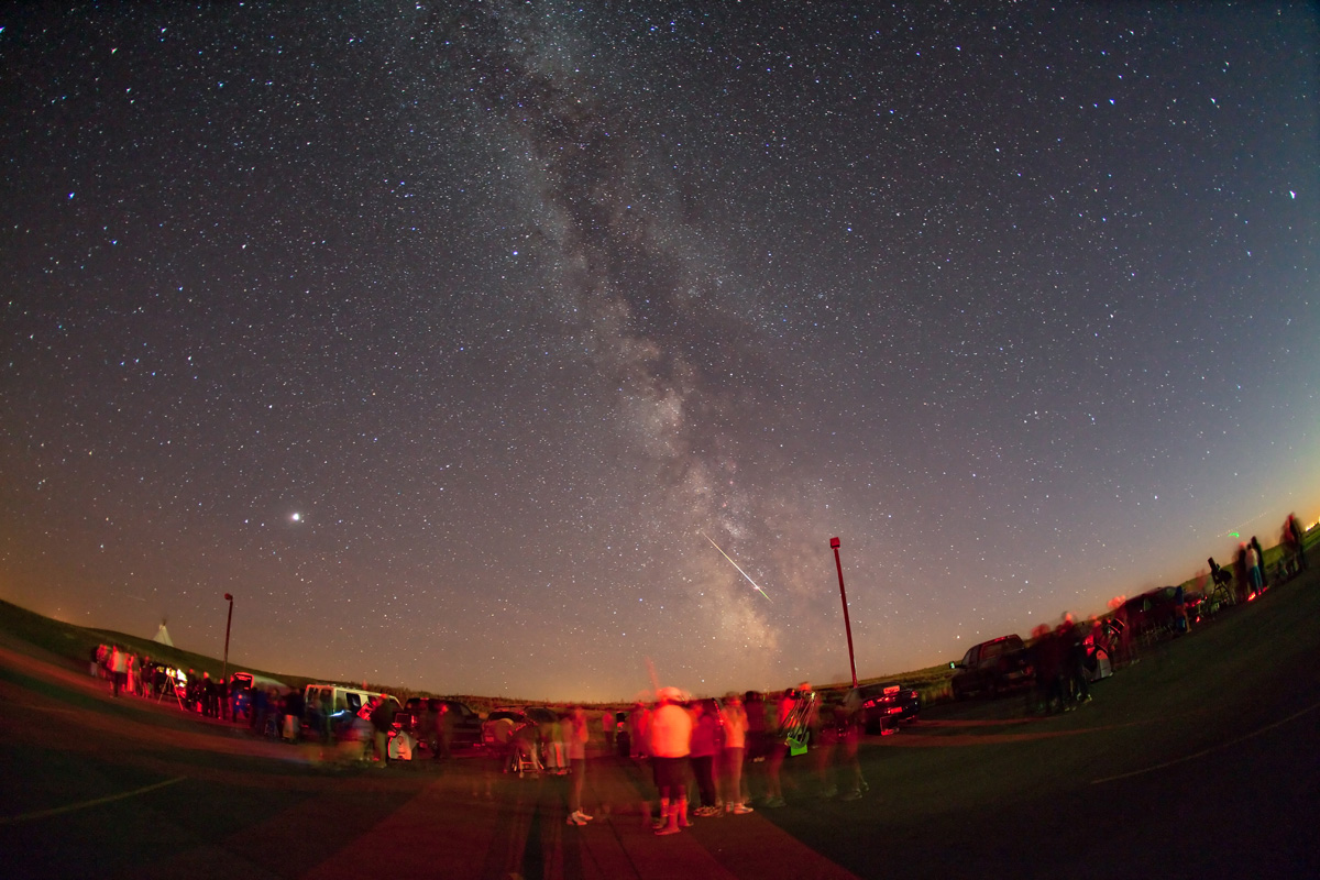 The Milky Way stretches above people, telescopes, and red observing lights at a rural star party in southern Alberta.