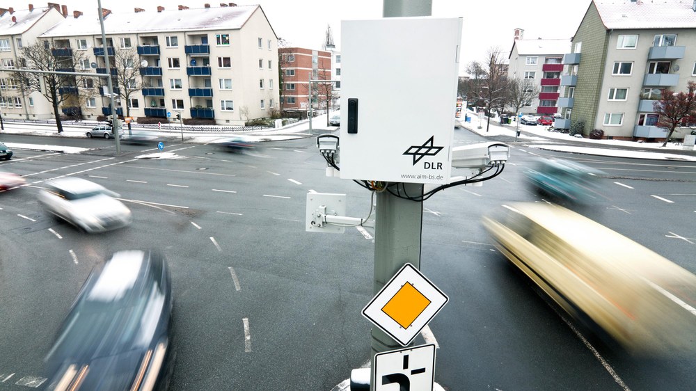 DLR monitoring equipment mounted above a snowy Braunschweig intersection, with cars blurred in motion below.