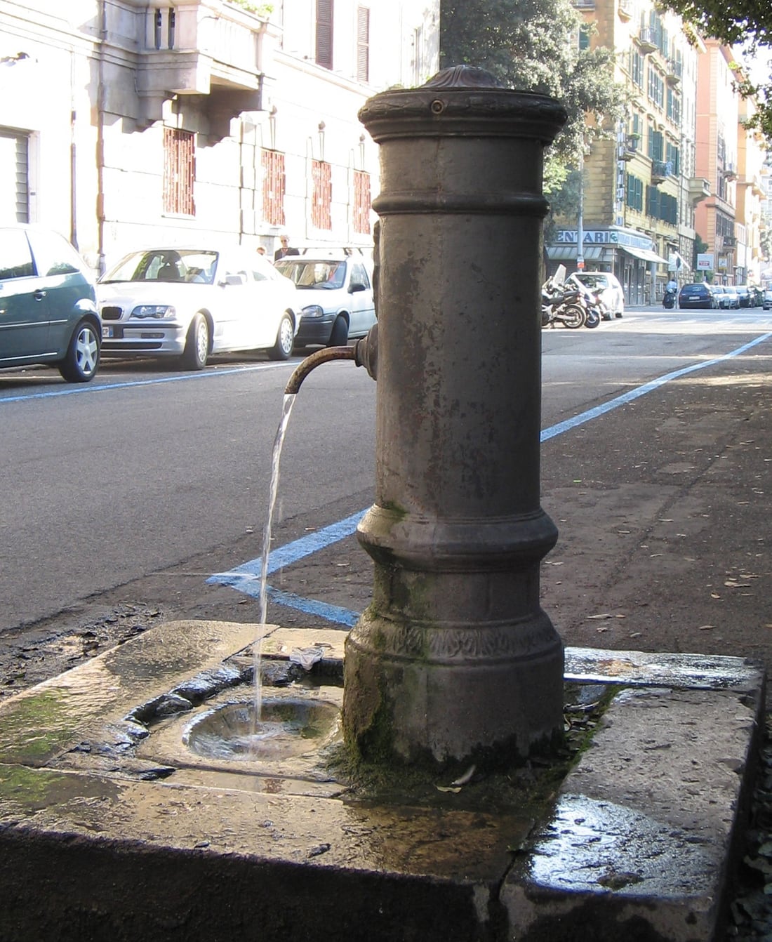 A cylindrical Roman nasone drinking fountain with a curved metal spout, standing on street paving.