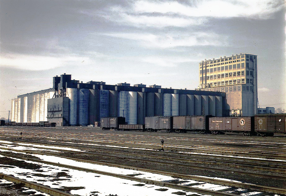 Concrete grain silos beside branching railroad tracks at the Santa Fe grain elevator in Kansas, photographed in color by Jack Delano.