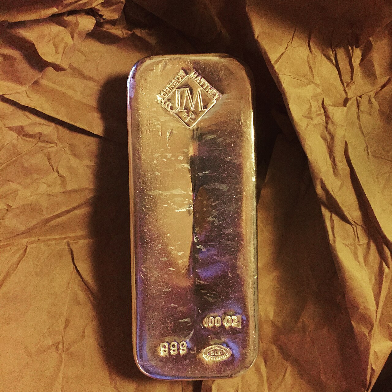A 100 troy ounce Johnson Matthey silver bullion bar photographed on a dark surface.