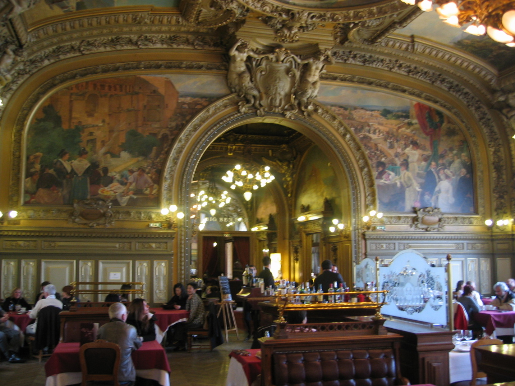 The ornate dining room of Le Train Bleu in Paris, with gilded moldings, painted ceilings, chandeliers, red banquettes, and set tables.