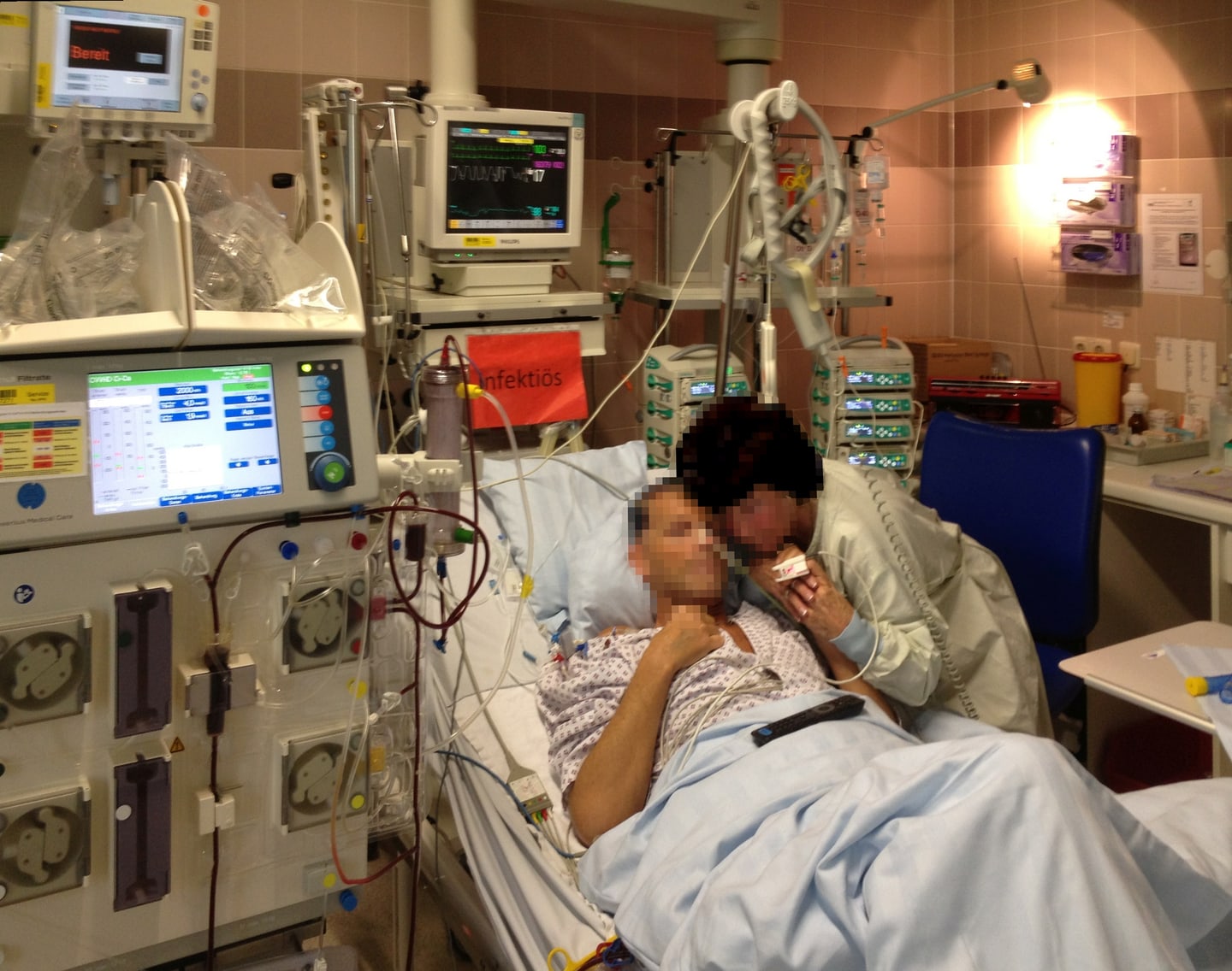 A patient and visitor in a German intensive-care room, surrounded by a bedside monitor, infusion pumps, and a dialysis machine; their faces are obscured.