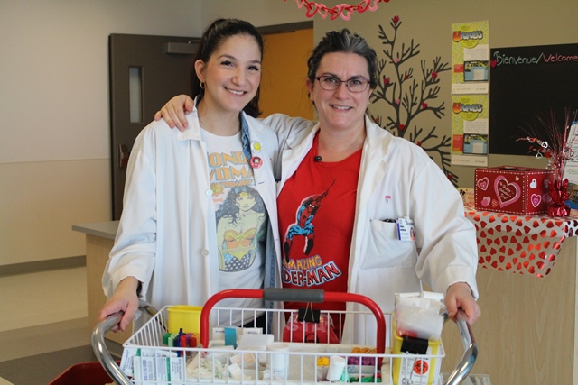 Two medical technologists in white coats and superhero T-shirts stand behind a specimen cart at Montreal Children's Hospital.