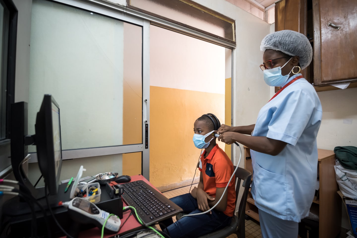 A clinician holds an ear probe at a seated patient's ear beside a computer in a hospital examination room.