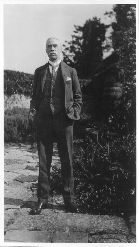 A. E. Housman stands on a stone garden path in a three-piece suit, with his hands in his pockets.